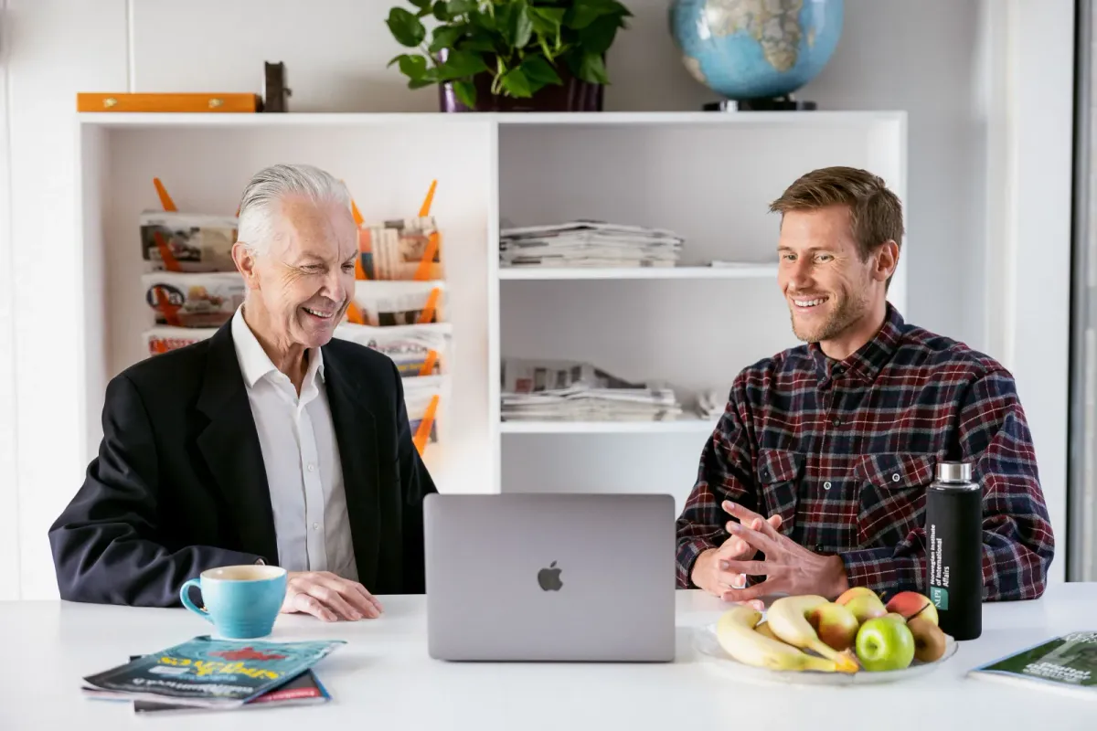Eldre mann i dress og yngre kollega i rutete skjorte i samtale ved bordet med laptop på lyst kontor