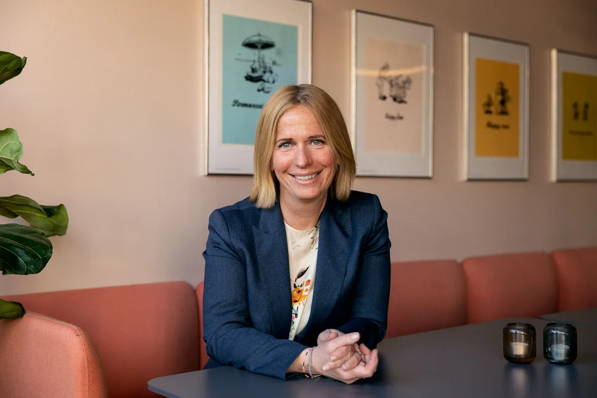 Smilende kvinne i marineblå blazer sitter ved et bord i en lun loungesone